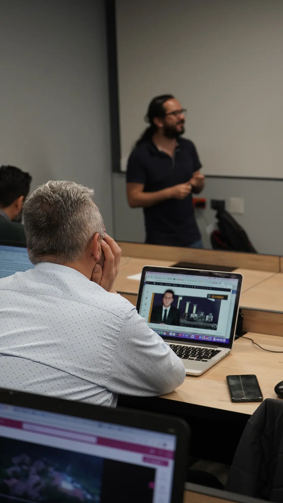 Un formador explica frente al grupo mientras un participante sigue la sesión en su portátil
